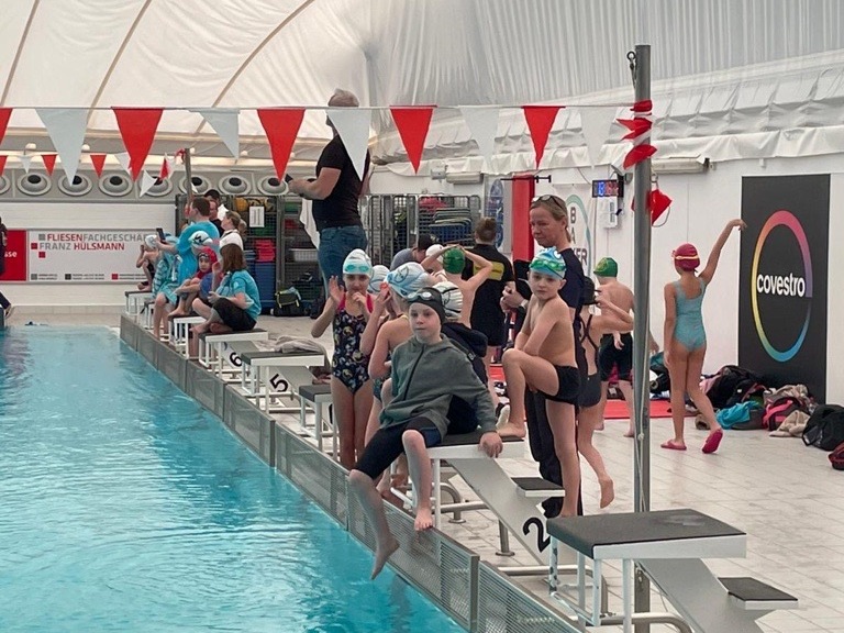 Kinder in Badekleidung warten auf den Sprung vom Startblock in einem Schwimmbad mit blauer Wasseroberfläche.