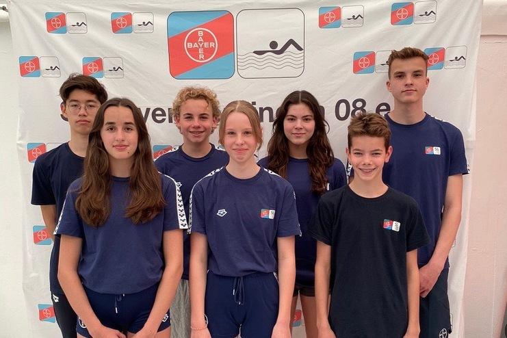Gruppe von sieben Jugendlichen in blauen T-Shirts vor einem Banner mit Schwimmmotiv und Vereinslogo.