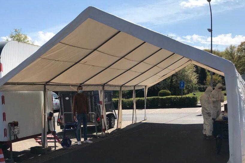 Zelt mit Personen in Schutzanzügen, ein Mann steht daneben; Beschilderung zur Unterstützung für ein Testzentrum.