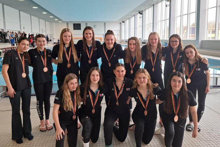 Gruppe von 15 Mädchen in schwarzen Shirts mit Medaillen, lächeln vor einem Schwimmbecken in einem Sportbad.