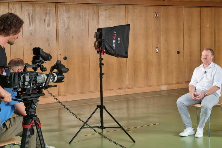 Drei Personen filmen ein Interview in einem Raum mit Holzverkleidung und einem Stuhl in der Mitte.
