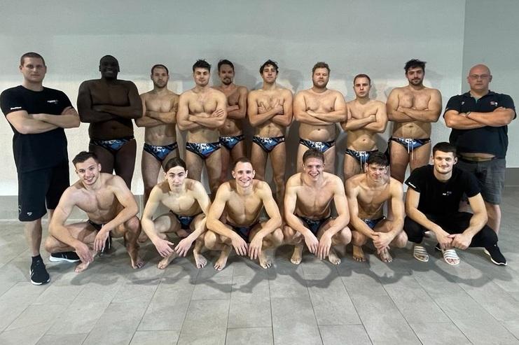 Gruppenfoto von 14 männlichen Wasserballspielern in Badehosen, stehend und sitzend, vor einer neutralen Wand.