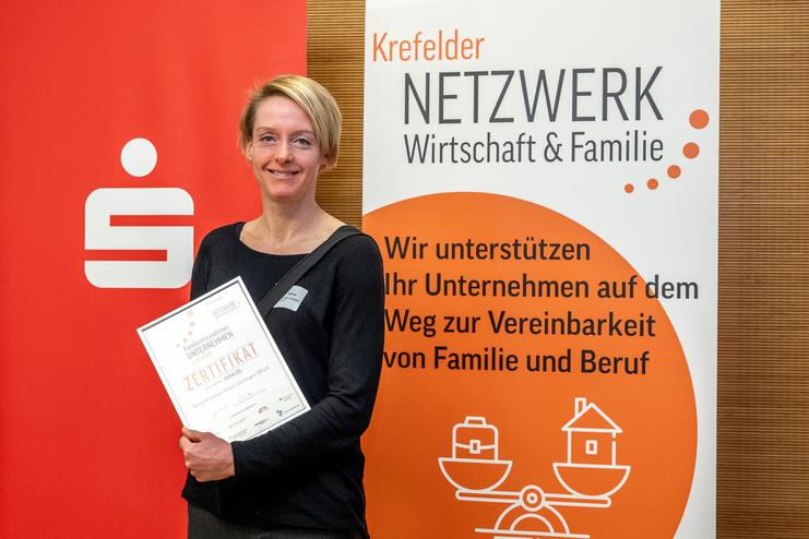 Frau mit Zertifikat in der Hand steht vor einem Banner des Krefelder Netzwerks für Wirtschaft und Familie.