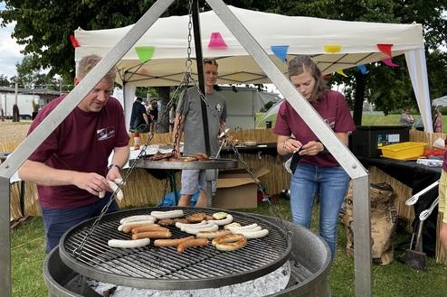Drei Personen grillen Würste auf einem offenen Grill unter einem Zelt mit bunten Fahnen im Hintergrund.
