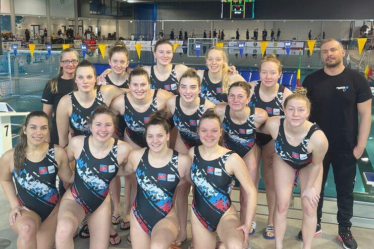 Gruppenfoto von 14 Schwimmerinnen in Wettkampfanzügen, posierend am Schwimmbecken mit Trainer im Hintergrund.
