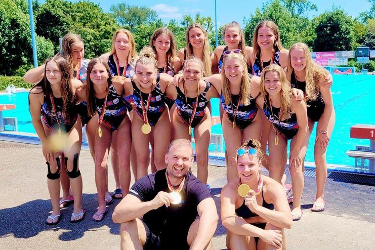 Gruppe von 13 Schwimmerinnen mit Medaillen, posed vor einem Schwimmbecken an einem sonnigen Tag.