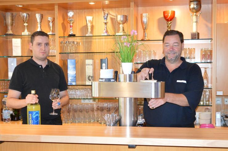 Zwei Bedienungen stehen an einer Bar, einer h&auml;lt eine Flasche, der andere zapft ein Getr&auml;nk. Im Hintergrund sind Pokale zu sehen.