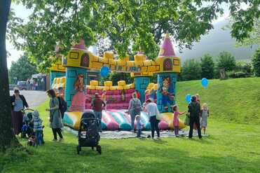 Gruppenspiel mit Kindern und Erwachsenen vor einer aufblasbaren Burg, umgeben von Wiese und Bäumen, im Freien.