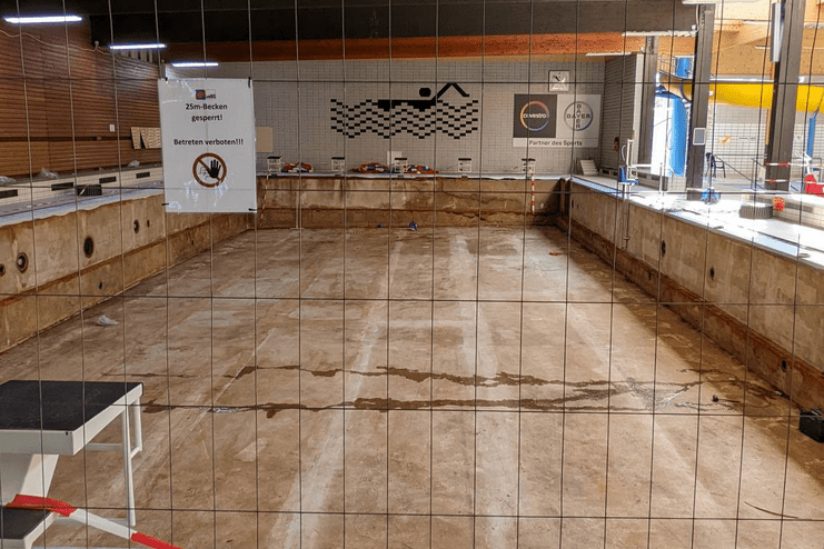 Leeres Schwimmbecken mit Absperrgitter und Hinweisschild "25m-Becken gesperrt" in einem Schwimmbad.