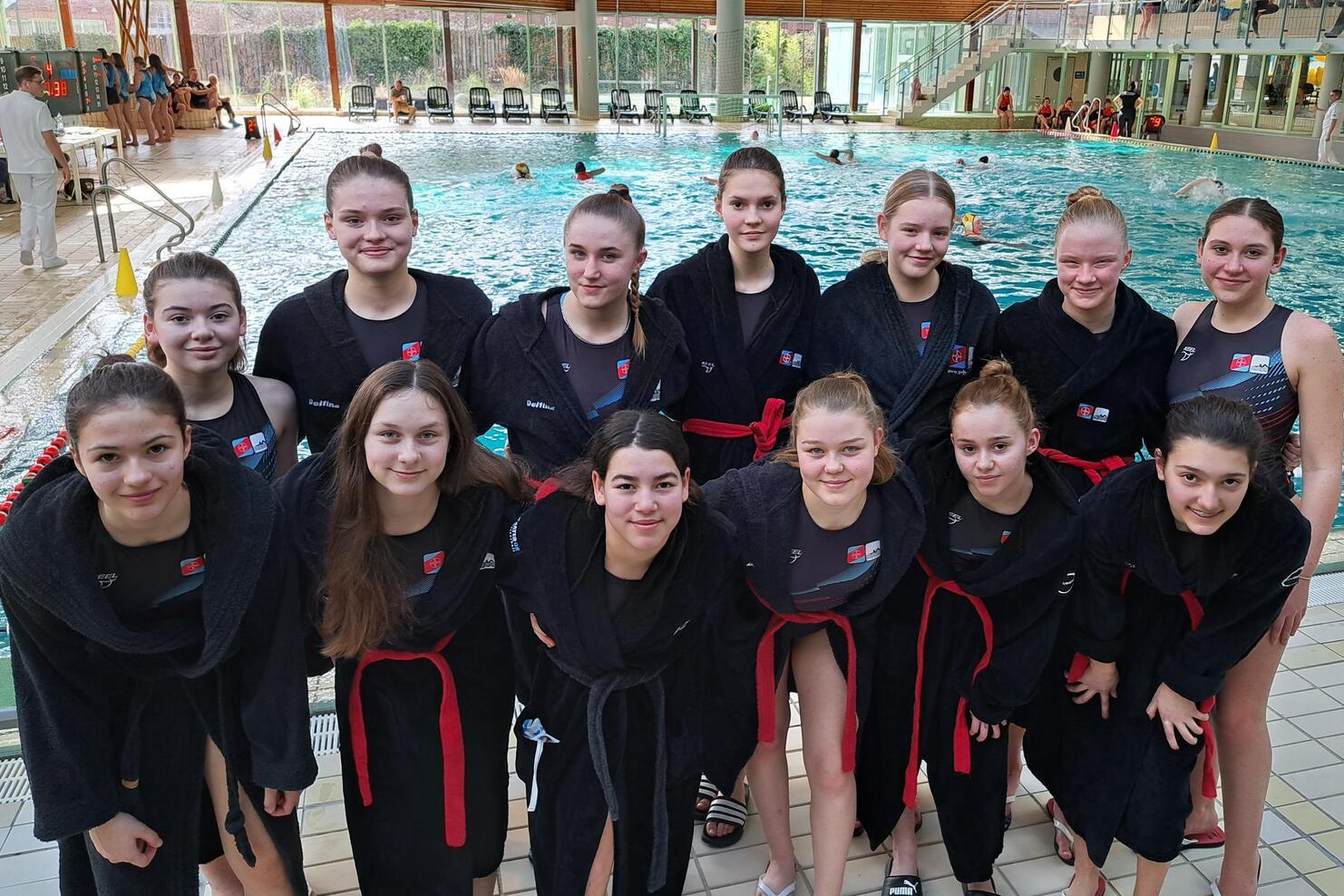 Gruppenumarmung von 14 jungen Schwimmerinnen in schwarzen Roben mit roten Gürtel am Beckenrand eines Schwimmbads.