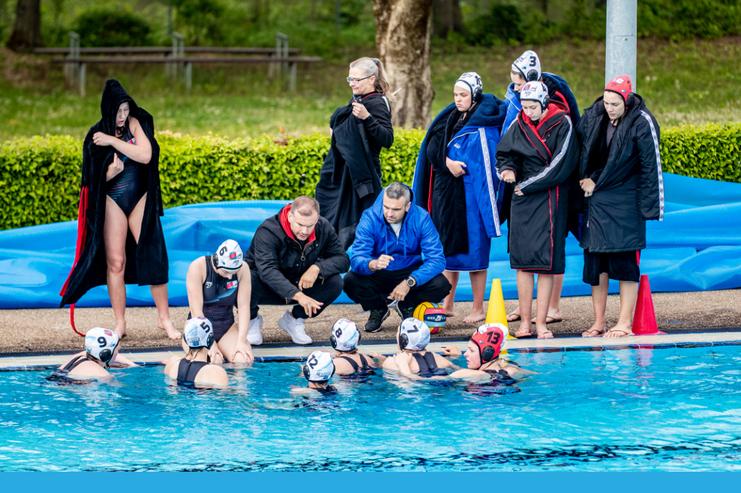 Wasserballmannschaft in Trainingsbekleidung um einen Pool, Spielerinnen besprechen Taktiken mit Trainern.
