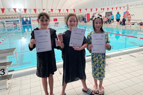 Drei Kinder in Badebekleidung stehen am Schwimmbad, halten Urkunden und freuen sich über ihre Erfolge.