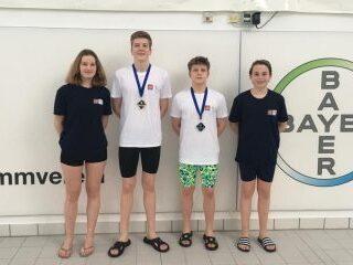 Vier Schwimmer stehen mit Medaillen um den Hals vor einer Wand mit dem Bayer-Logo und einem Schwimmvereinsymbol.