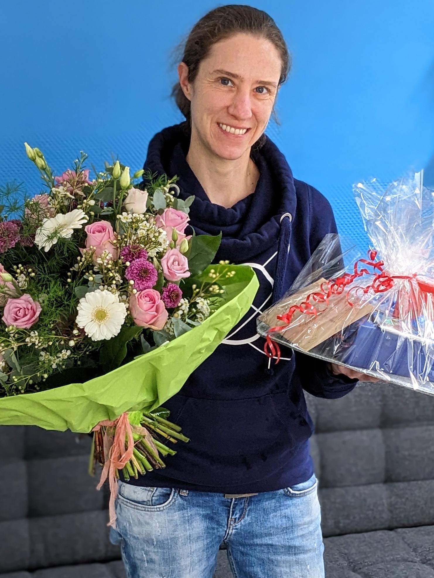 Frau mit einem gro&szlig;en Blumenstrau&szlig; und einem Geschenk in der Hand steht vor einer blauen Wand, auf einem grauen Sofa.