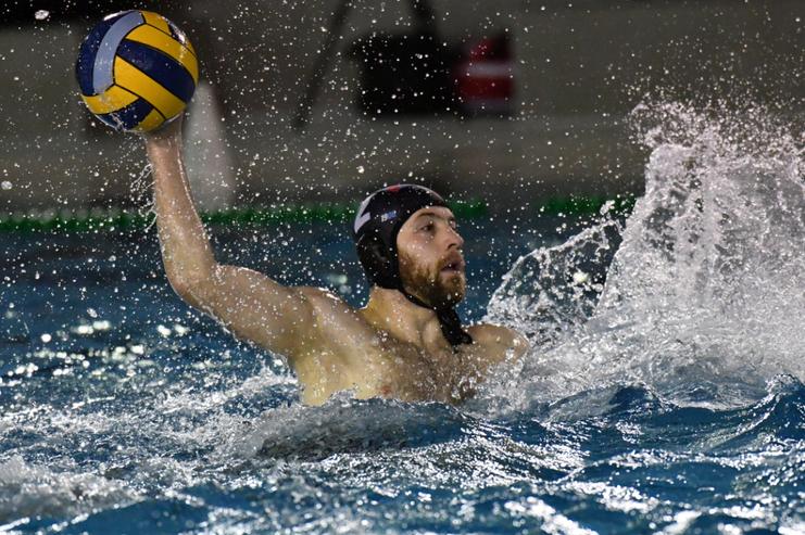 Ein Wasserballspieler mit einer gelb-blauen Kugel in der Hand spritzt Wasser beim Wurf in einem Schwimmbecken.
