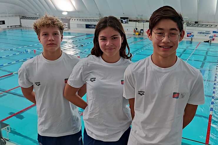 Drei Jugendliche in weißen T-Shirts stehen vor einem Schwimmbecken mit blauer Wasseroberfläche und roten Fähnchen.