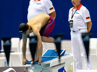 Wettkampfschwimmer auf dem Startblock, bereit zum Sprung, während Trainer im Hintergrund beobachtet.