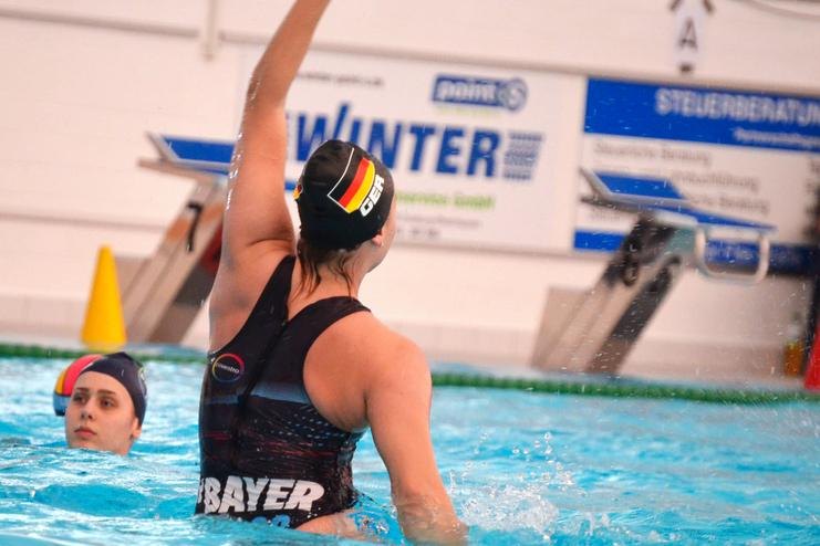 Zwei Wasserballspielerinnen im Becken, eine hebt den Arm, während die andere im Hintergrund sitzt.