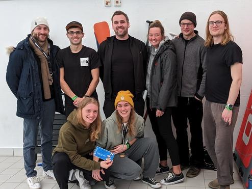 Gruppe junger Menschen posiert l&auml;chelnd vor einer Wand mit roten Surfboards und einer grauen Sportausr&uuml;stung.