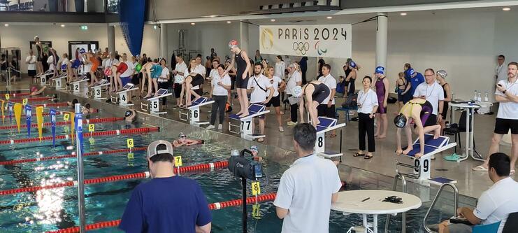 Schwimmerinnen und Schwimmer stehen auf Startblöcken an einem Wettkampfbecken, Zuschauer und Trainer beobachten die Veranstaltung.