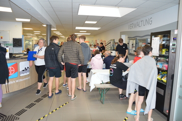 Menschen stehen in einem Schwimmbadbereich, einige kaufen Snacks, andere warten oder unterhalten sich.