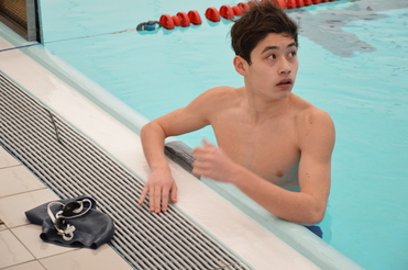 Junger Schwimmer sitzt am Beckenrand, schaut überrascht und hat eine Schwimmkappe sowie Schwimmbrille neben sich.