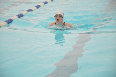 Junge schwimmt im Becken, trägt Badekappe und Schwimmbrille, Wasser spritzt um sie herum.
