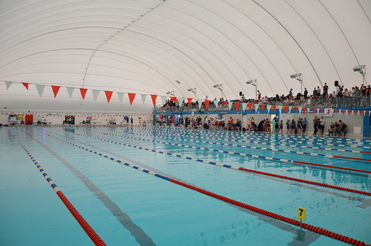 Schwimmhalle mit mehreren Bahnen und Rot-Weißen Wimpeln, Zuschauern auf der Tribüne und einer hohen Kuppeldecke.