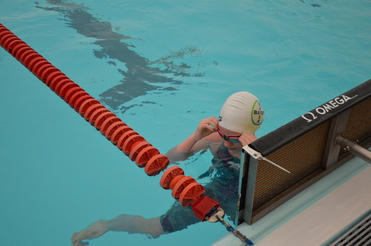 Schwimmerin in Badekleidung setzt ihre Schwimmbrille an der Wand einer Schwimmhalle auf. Schwimmbahnmarkierungen im Wasser.