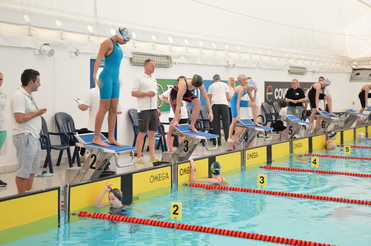 Schwimmerinnen stehen startbereit auf den Startblöcken neben dem Bassin, während Trainer und Zuschauer beobachten.