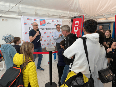 Gruppierung von Menschen bei einer Veranstaltung, während ein Redner auf einer Bühne spricht, im Hintergrund eine Bannerwerbung.