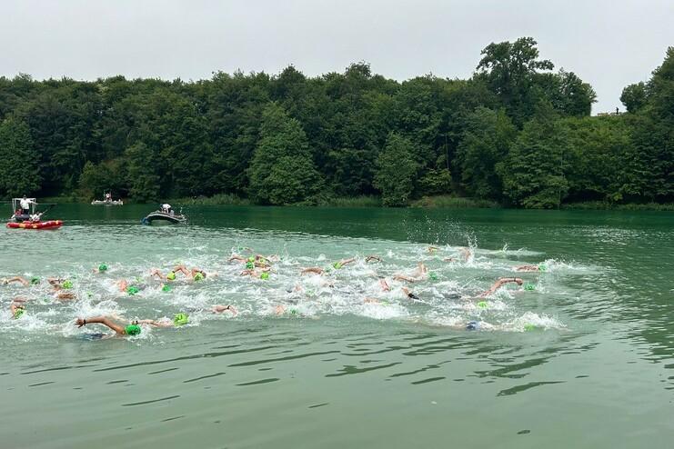 Schwimmer in Neon-Kappen durchqueren einen grünen See, umgeben von Bäumen und Booten.