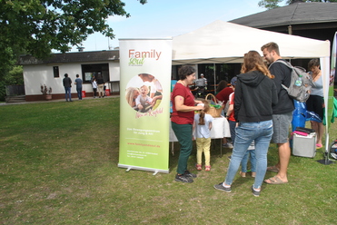 Menschen stehen vor einem Infostand mit Banner in einem Park, umgeben von Bäumen und einem Zelt im Hintergrund.