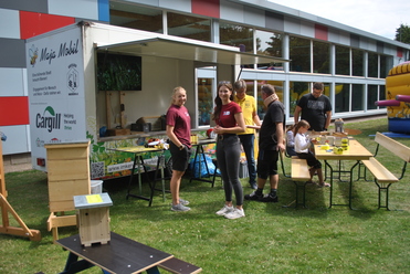 Zwei Frauen und mehrere Erwachsene stehen vor einem mobilen Imkerstand mit Bienenkästen und Bastelmaterial auf Wiesenfläche.