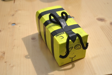 Gelbes Geschenkpaket mit schwarzem Band und Deko aus Pfeifenreinigern auf einem Holztablett.