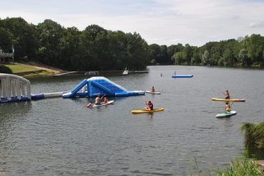 Kinder paddeln auf Stand-Up-Paddleboards in einem See, mit einer aufblasbaren Spiellandschaft im Hintergrund.