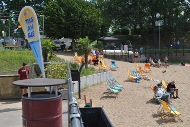 Strandbereich mit Liegestühlen, einem Banner „Tag am See“ und Gästen, die sich am Sand entspannen.