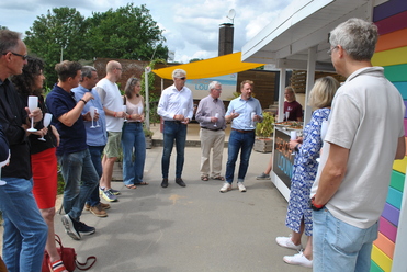 Gruppe von Menschen steht im Freien vor einem bunten Bereich, während ein Mann mit Mikrofon spricht.