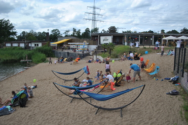 Menschen entspannen am Sandstrand eines Sees, umgeben von Liegen, Hängematten und einer bunten Strandbar.