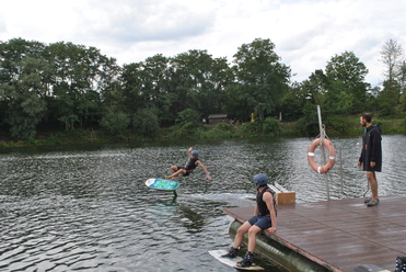 Ein Wassersportler springt mit einem Wakeboard über das Wasser, während zwei Zuschauer auf einem Steg sitzen.