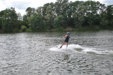Person auf einem Wakeboard fährt über einen See, hinterlässt Wellen, umgeben von Bäumen und einer bewölkten Himmel.