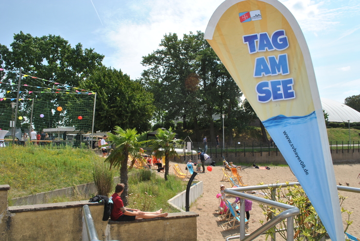 Fahne mit der Aufschrift "TAG AM SEE" neben einer Sandfläche mit Liegestühlen und Menschen.
