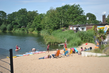 Blick auf einen Sandstrand am See mit Sonnenliegen und Personen beim Paddeln und Spielen. Grüne Ufervegetation im Hintergrund.