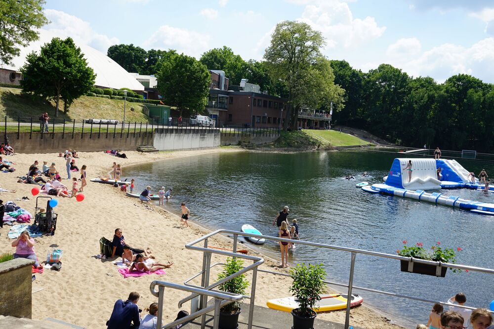 Strand mit Badegästen, Sonnendecken und einem aufblasbaren Spielgerät im Wasser, umgeben von Bäumen und Gebäuden.