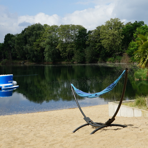 Hängematte auf einem Sandstrand am Wasser, umgeben von Bäumen und blauem Himmel mit Wolken.