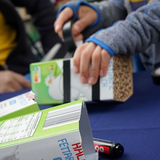 Kinderhände befestigen Milchkartons mit Klebeband auf einem Tisch, um ein Bastelprojekt durchzuführen.