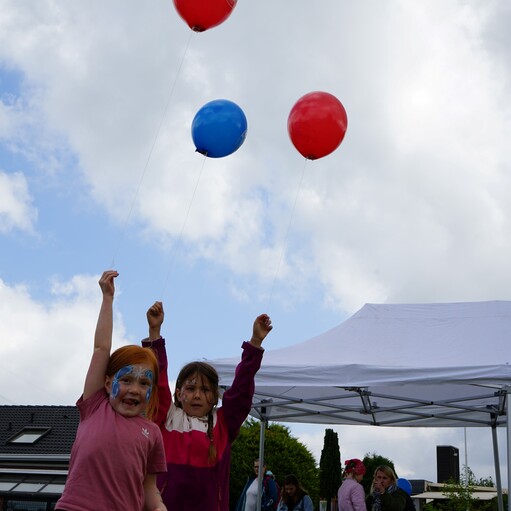 Zwei Mädchen halten strahlend rote und blaue Ballons, unter einem Zelt stehen weitere Personen im Hintergrund.