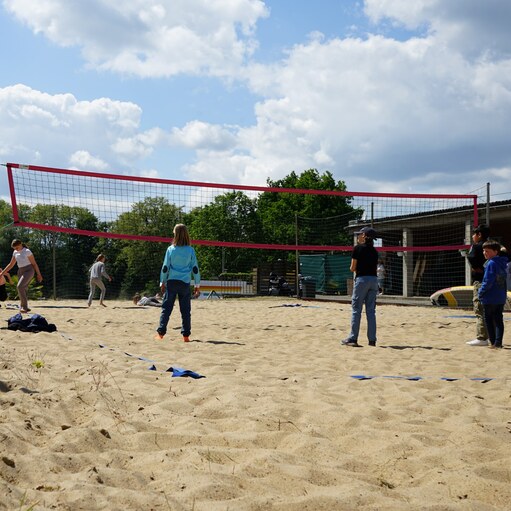 Gruppe von Kindern und Jugendlichen spielt Volleyball auf einem Sandplatz unter einem wolkigen Himmel.