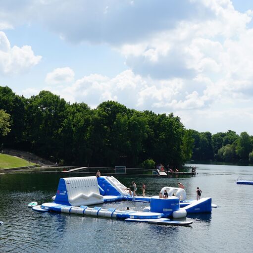 Blick auf einen Badesee mit Wasserspielelementen, Schwimmern und Menschen am Strand bei schönem Wetter.