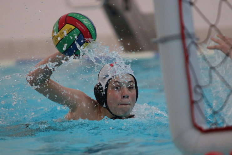 Wasserball-Spieler wirft einen bunten Ball, umgeben von Spritzwasser, vor einem Tor im Becken.